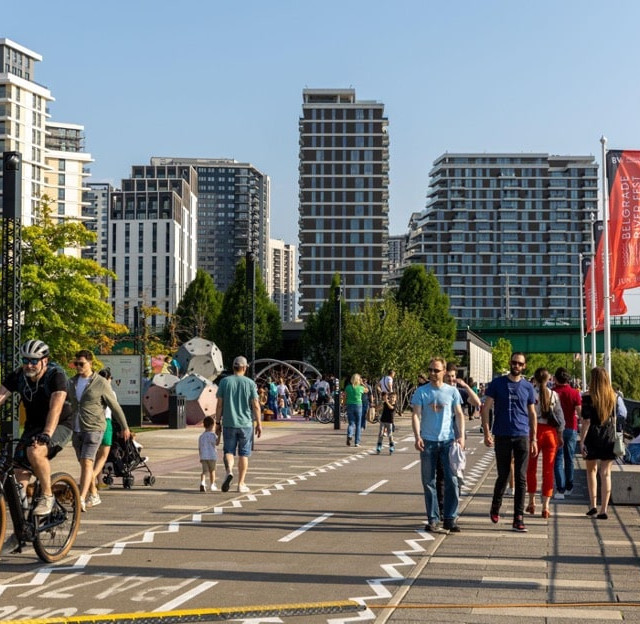 Ljudi šetaju i voze bicikle, a deca se igraju na igralištu na Sava Promenadi tokom sunčanog, letnjeg dana.